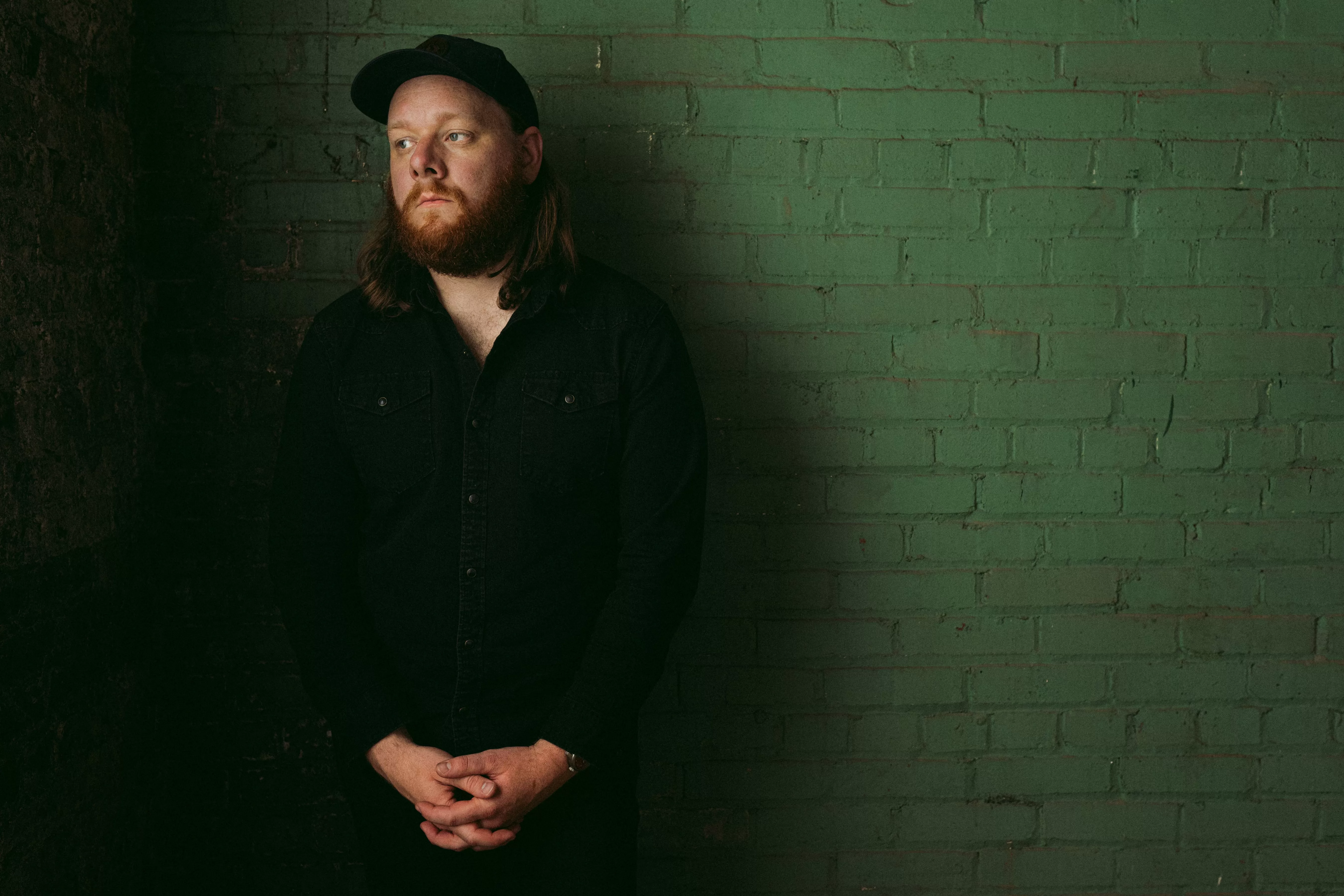 Mountain Thief with long auburn hair and beard wearing black, standing against green brick wall