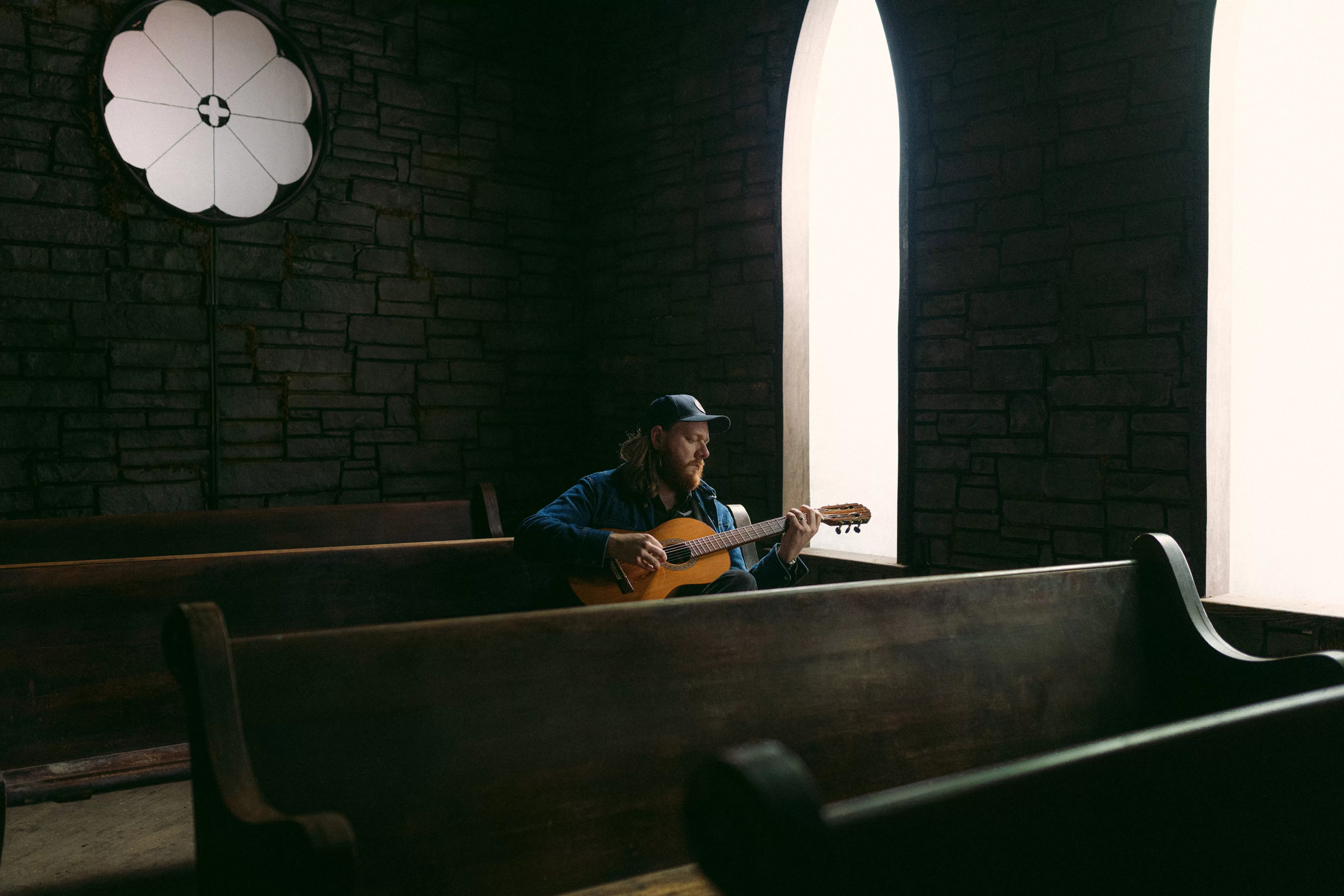 Focused Mountain Thief playing acoustic guitar on church pew with arched windows in background